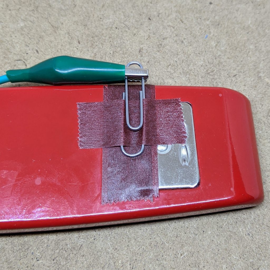 stapler anvil with tape across it supporting a paperclip, taped down. A green alligator clip is attached to the free end of the alligator clip