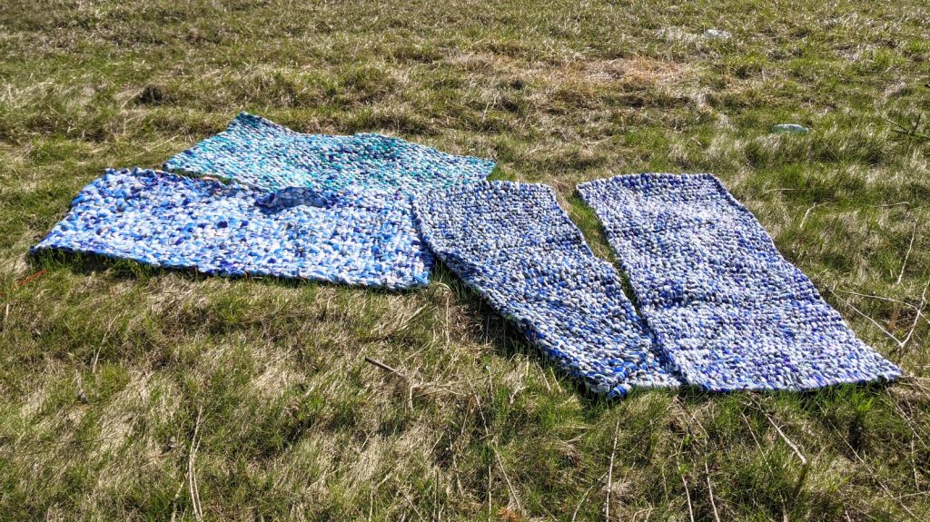 blankets woven from hundreds of milk outer bags: dark blue, light blue, white, yellow. Mats are laid out on tussocky grass