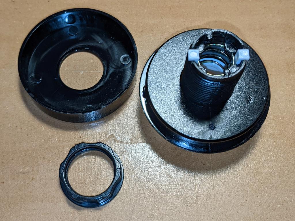 the top of the button disassembled into its main parts: bezel ring at top left, threaded lock ring at bottom, and main button mechanism. The mechanism is upside down, so the return spring and button actuators can be seen inside the threaded shaft