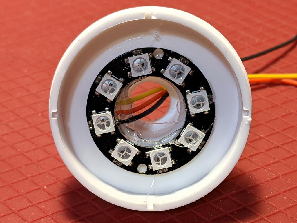 White button top underside with an 8 RGB LED ring hot glued into it. Three thin insulated wires (from top: yellow (data), red (5 V) and black (GND)) are previously soldered behind the LED board, and are secured against strain with a large deposit of hot glue