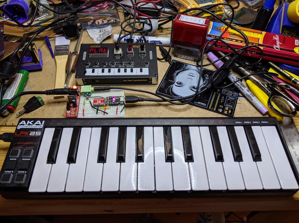 on a messy desk, a small USB midi keyboard is connected to a Korg NTS-1 mini synthesizer via a small micro-controller board that acts as a USB host for the Akai keyboard, converting USB MIDI to traditional MIDI for the Korg