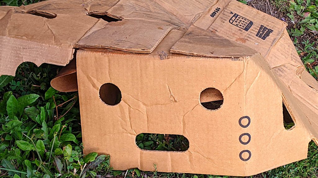 a broken up brown cardboard produce box lies upside down, with two circular vents suggesting eyes and a flat oval carrying handle slot suggestion a distraught mouth. On the corner edge, leading down from the "eye" on the right, are three printed circles like teardrops