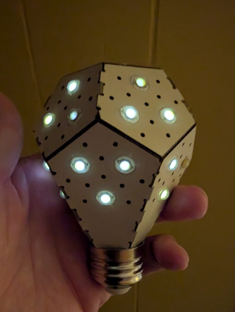 A hand holds an oddly-shaped light-bulb. It is a regular dodecahedron (12 sides) but with five of the lower faces extended into kite shapes. The top six faces are regular oentagons. There are three white LEDs on 11 of the sides. All but one of the LEDs are glowing, despite the Edison Screw mount not being inserted into an outlet